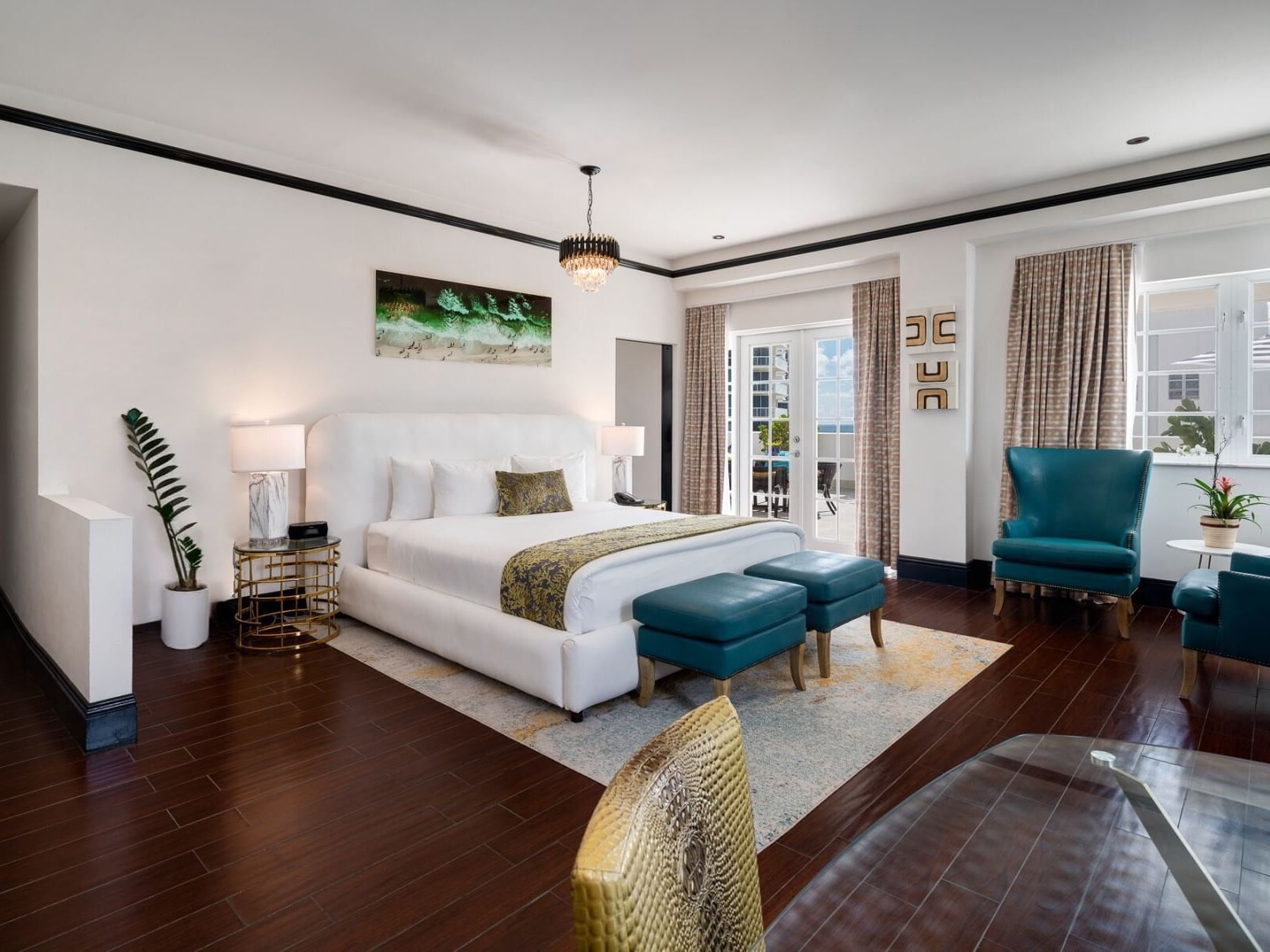 Elegant bedroom with ocean views, large bed, two ottomans, nightstands, chandelier, and teal chairs near a glass table.