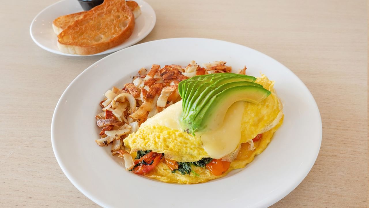 Plate with omelet, avocado, and hash browns next to plate with toast.