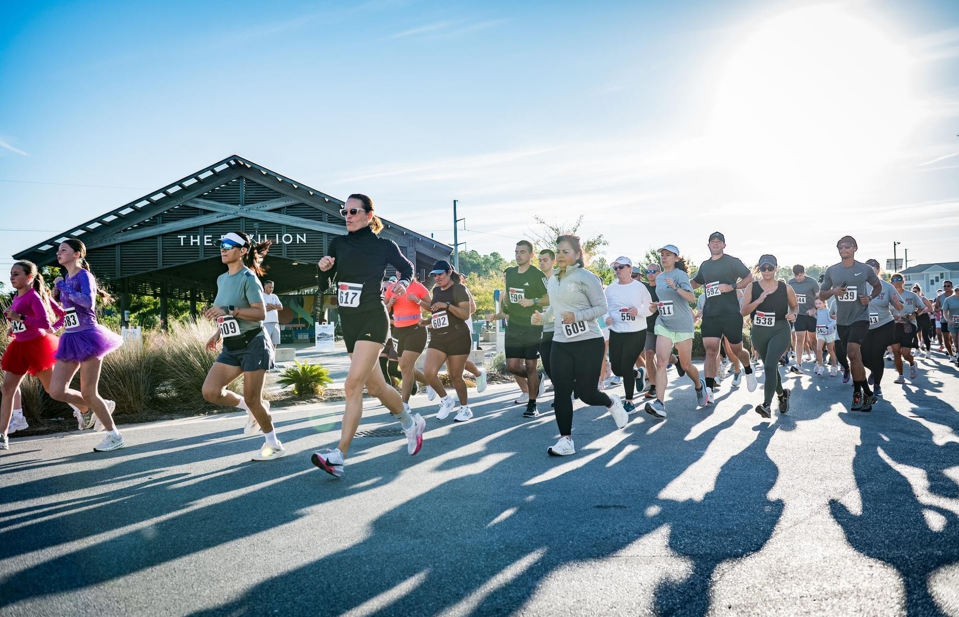 Runners wearing numbered bibs race past a covered pavilion under a blue sky at Watersound Town Center.