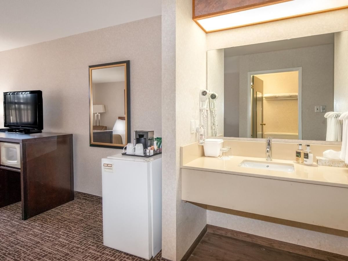 A hotel room with a bathroom, sink, mirror, small fridge, TV, and dresser.