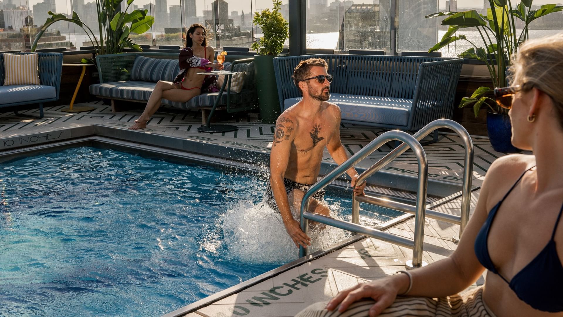 Guests enjoying a rooftop pool with city skyline views, lounge seating, and tropical plants in urban setting