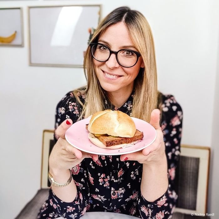 Woman with blonde hair holds a pink plate with a sandwich in front of her.