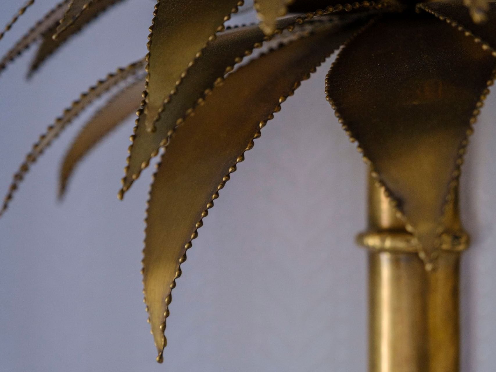 Detailed close-up of a golden palm leaf lamp with a central golden stem against a light blue background.