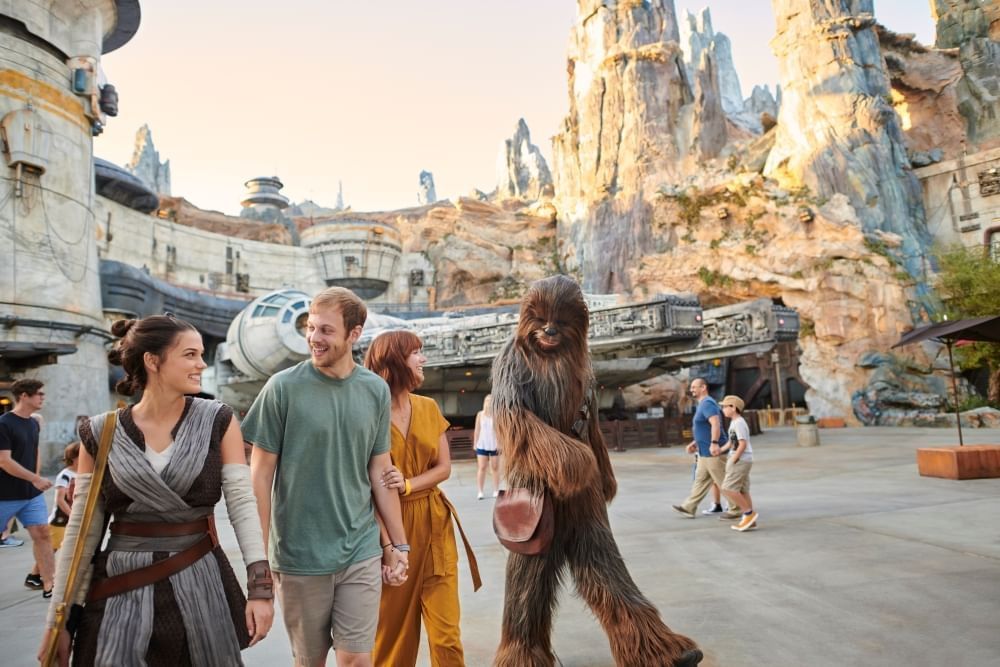 A man and woman walk through a rocky Star Wars environment in front of the Millennium Falcon with Chewbacca and a Jedi woman in robes.  