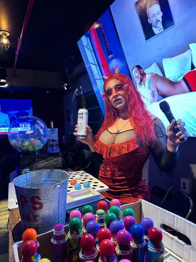 A performer in a red dress holds a microphone, surrounded by colorful game pieces in a lively venue at The Gaythering