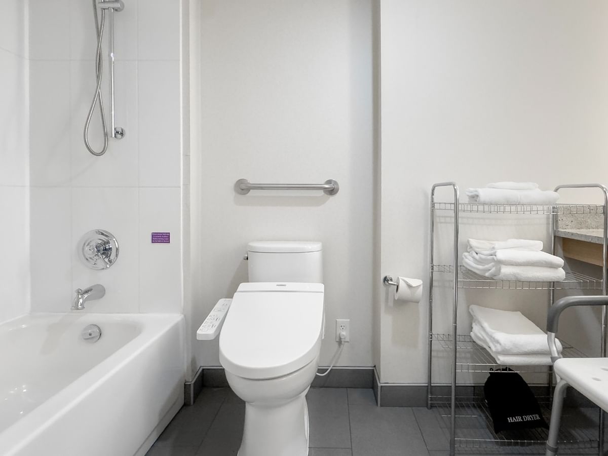 An accessible bathroom with a white bathtub, toilet, grab bars, shower, and folded towels on a shelf.