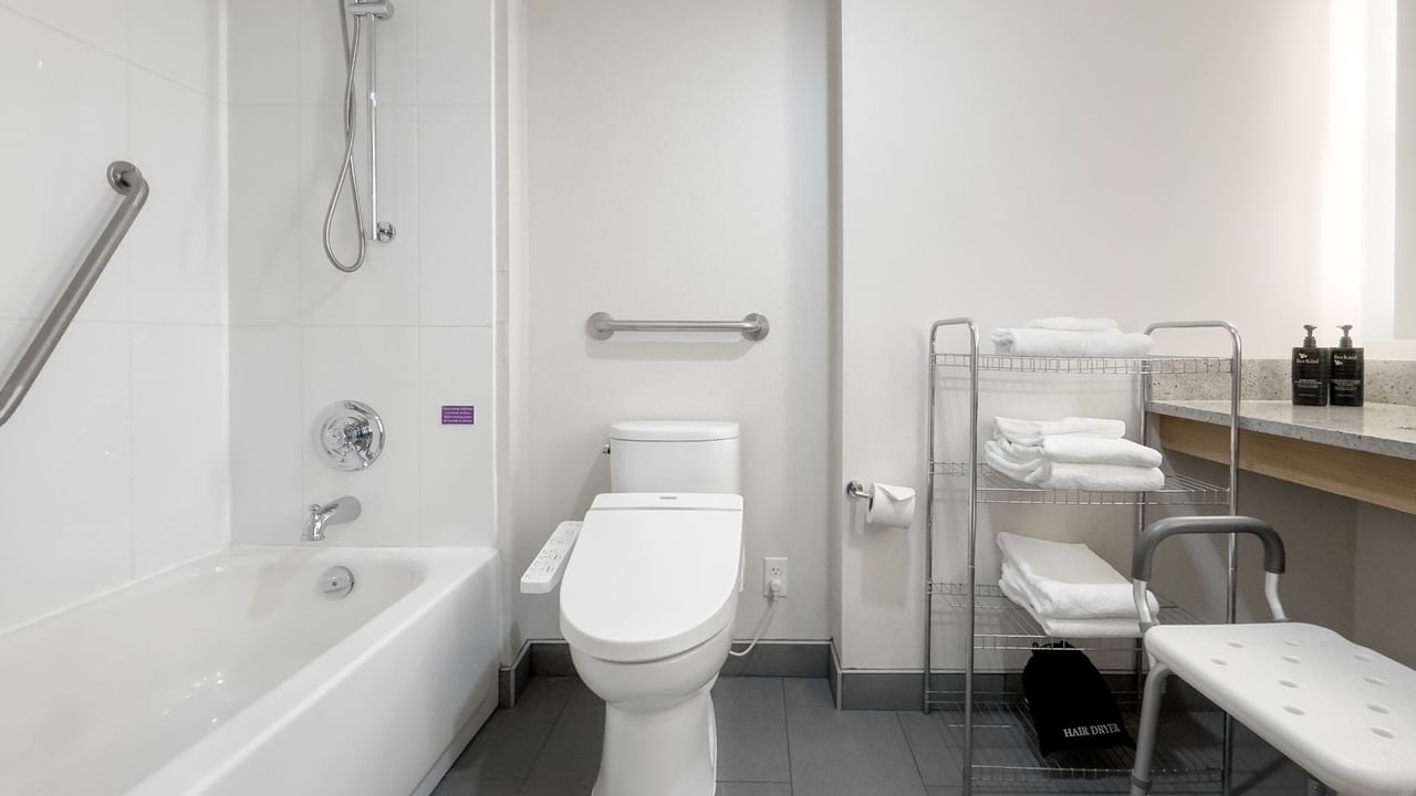 An accessible bathroom with a white bathtub, toilet, grab bars, shower, and folded towels on a shelf.