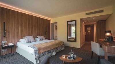 A hotel room with a bed, pillows, wooden table, chair, desk, mirror, and carpeted floor.