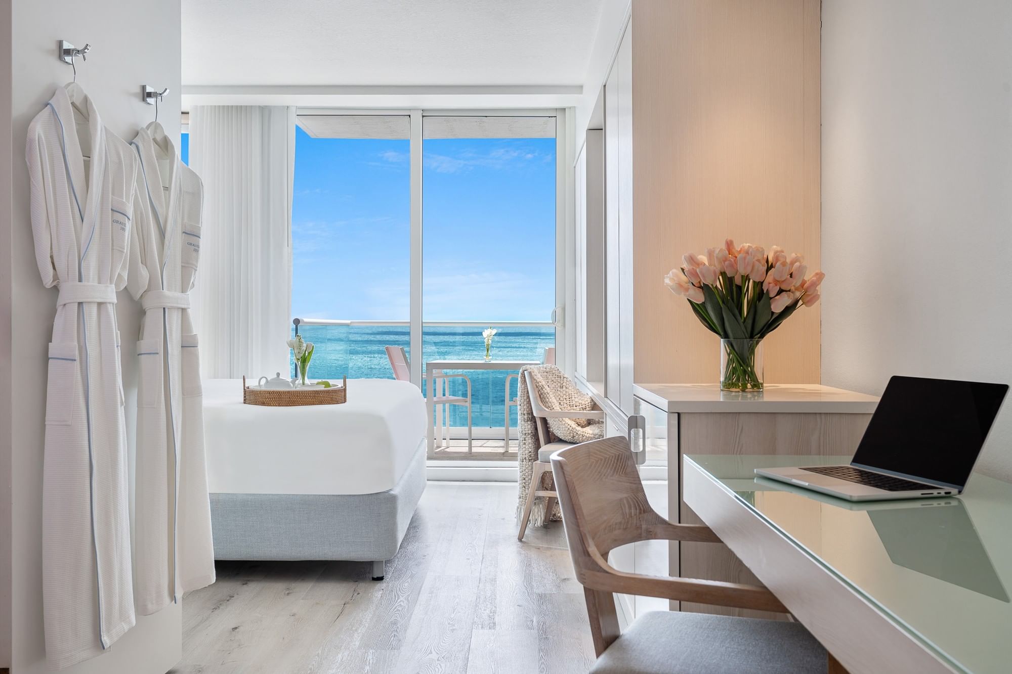 A hotel room with a white bed, desk, flowers, and robes, overlooking the ocean.
