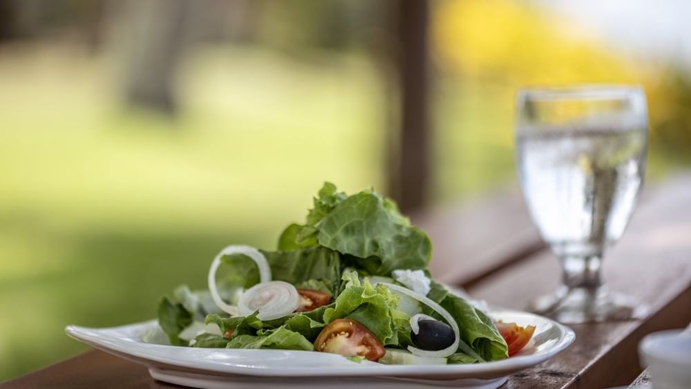 Fresh green salad with tomatoes & olives on a white plate by a water glass in Tambua Restaurant at Tambua Sands Beach Resort