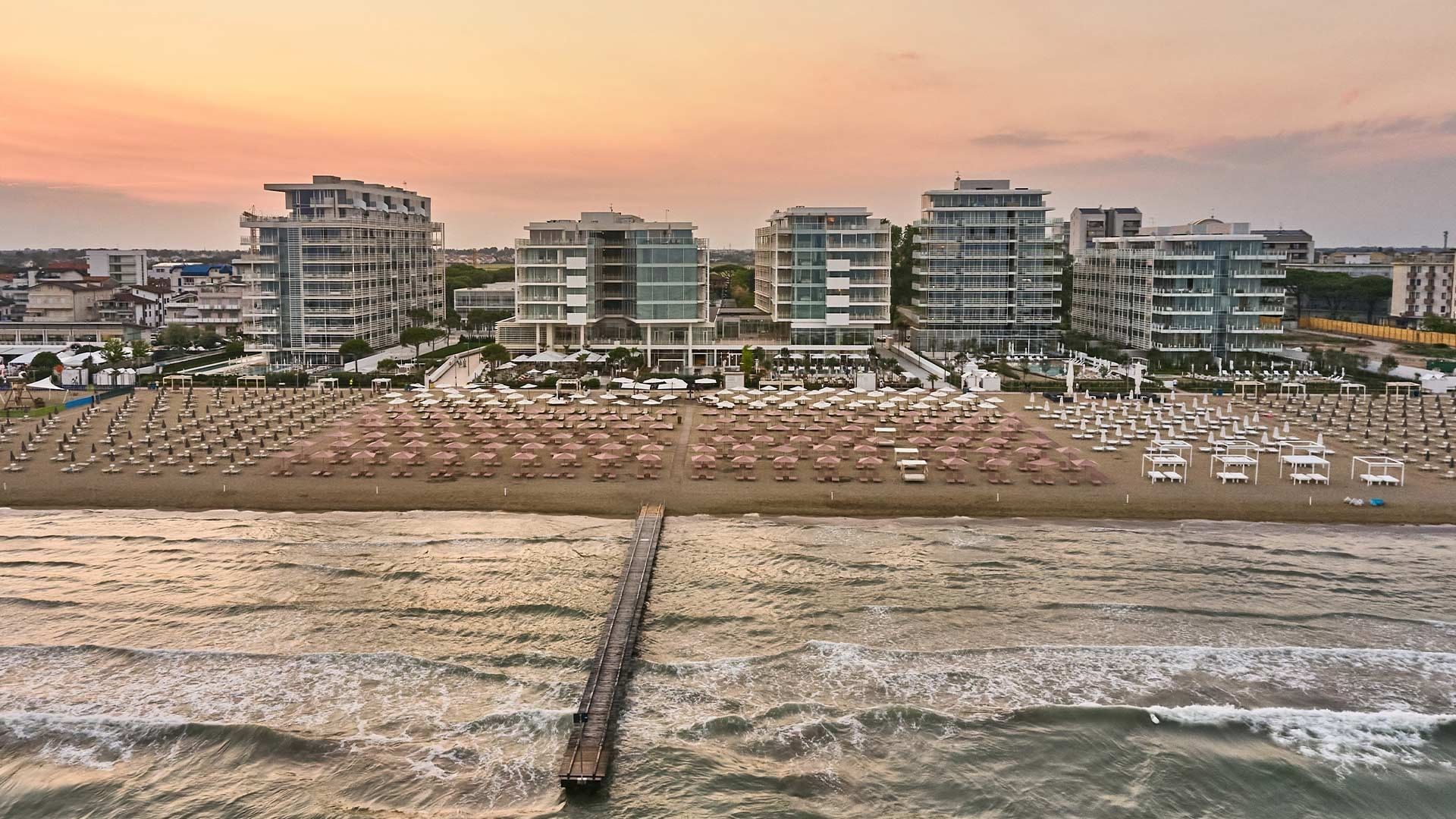 Aerial view of a resort with beach, lounge chairs, umbrellas, and buildings at sunset.