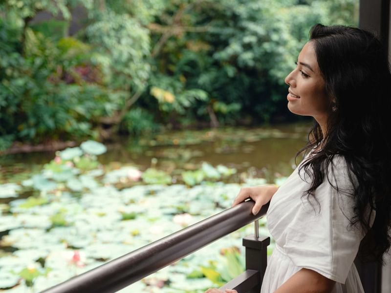 Malaysian woman enjoying peaceful moment on balcony overlooking serene pond with lily pads and lush greenery at The Banjaran Hotsprings Retreat