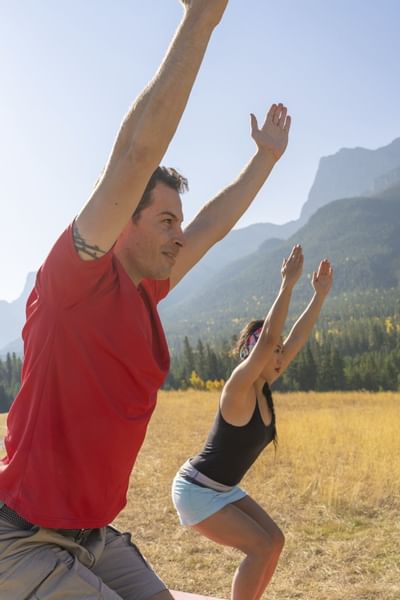 Two individuals doing yoga outdoors in a grassy field, surrounded by mountains near Blackstone Mountain Lodge