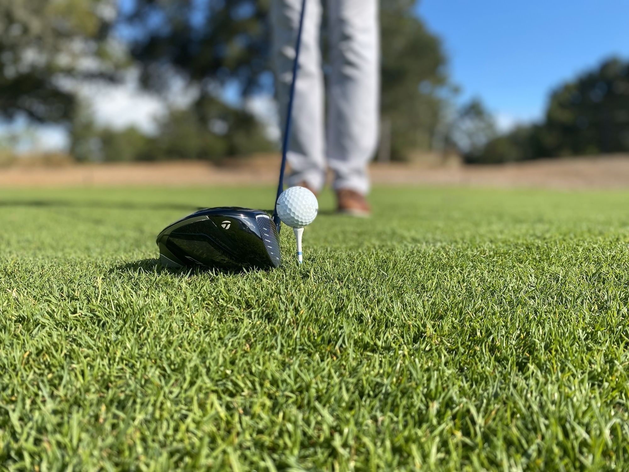 Golfer in khaki pants and brown shoes preparing to hit a golf ball on a green field.