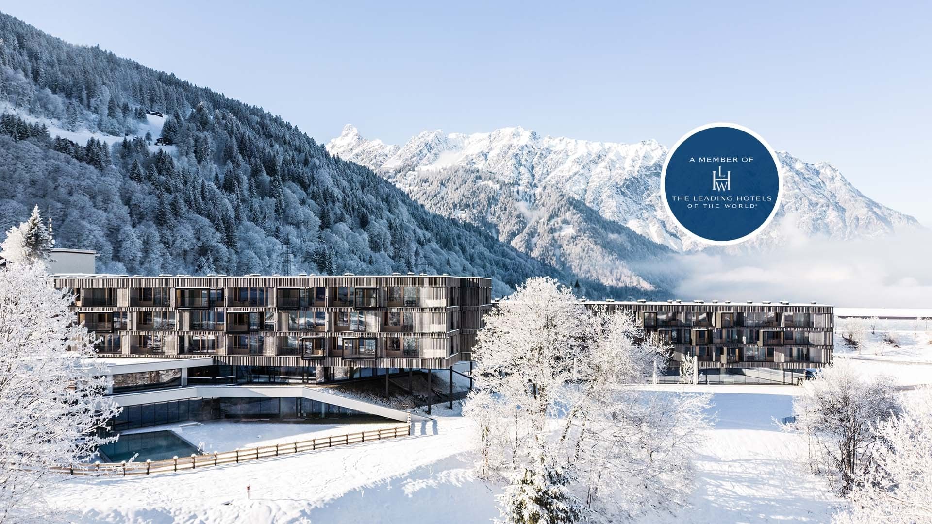 Modernes Hotelgebäude in den verschneiten Alpen, umgeben von schneebedeckten Bäumen und Bergen.