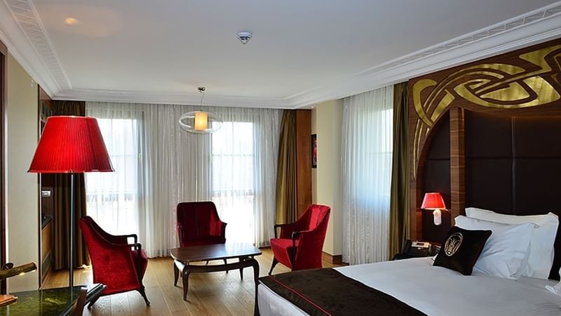 Red chairs by a coffee table surrounding a plush bed under a gold headboard in Junior Suite at Warwick Ankara