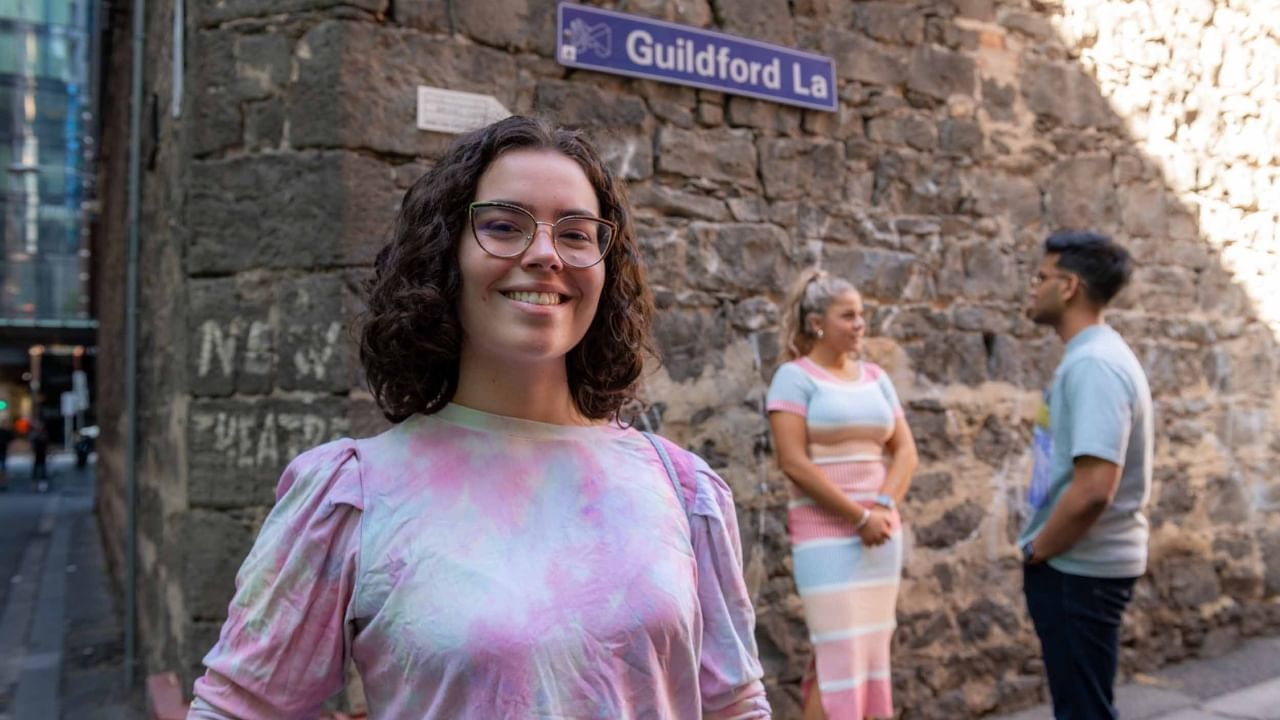 Smiling resident stands in front of a stone wall in Melbourne laneway with friends standing behind her talking. 
