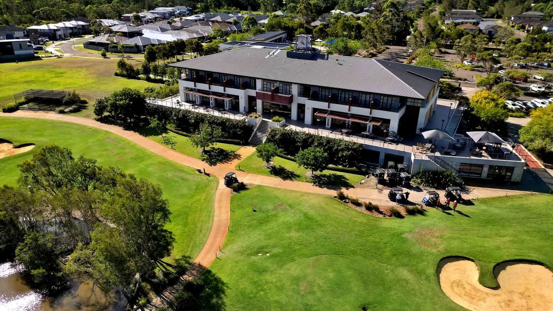 Aerial view of a golf course with a large club house, a pond, and several buildings in the background.