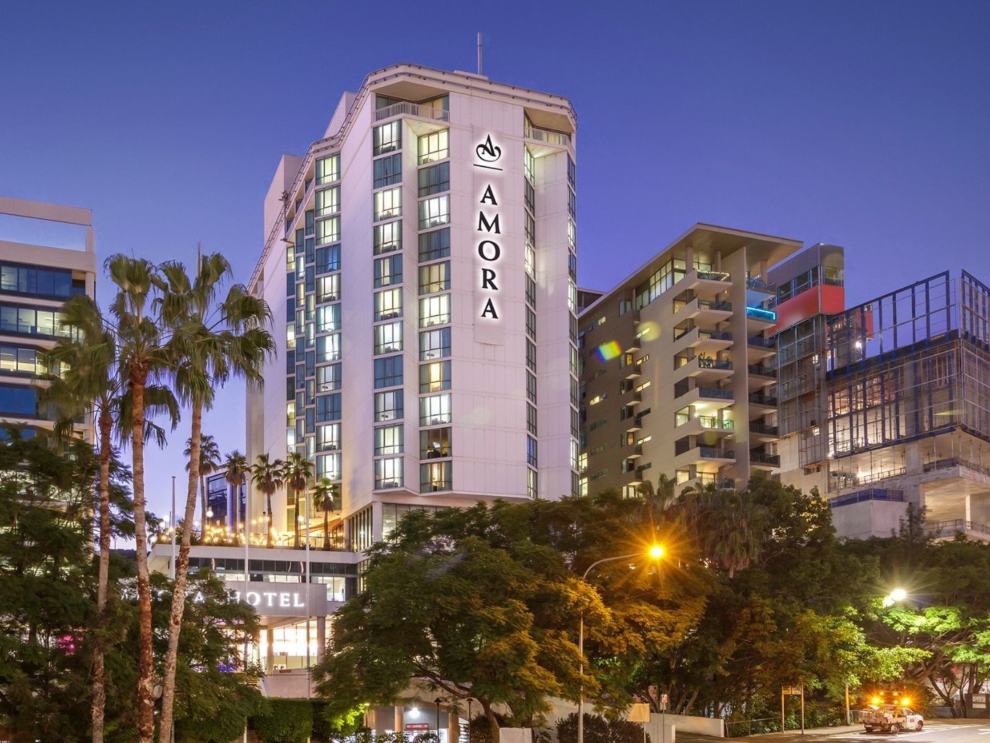 Exterior view of the tall white hotel building at dusk, featuring the logo of Amora Brisbane Hotel