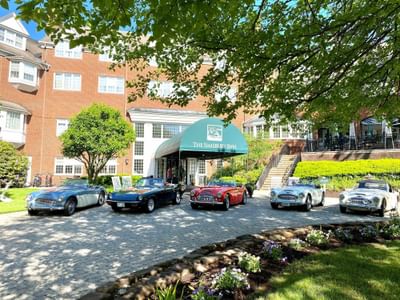 The Simsbury Inn entrance driveway features a lineup of classic, colorful convertible sports cars parked on the pavers