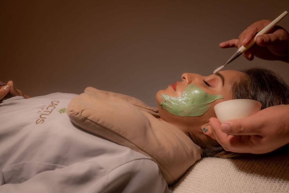 Una persona está acostada con una mascarilla facial verde, envuelta en una toalla.