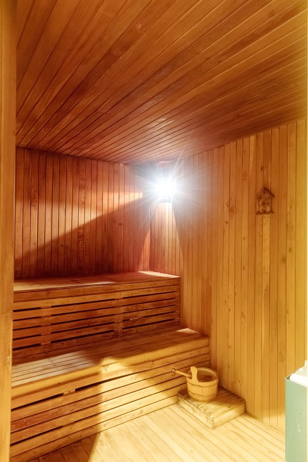 Tiered wooden benches in a classic sauna room under a bright, glowing light by a bucket at Kampala Serena Hotel