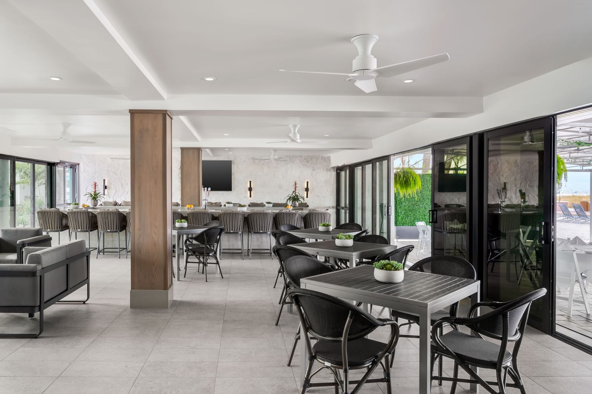 A modern dining area with tables and chairs, a bar in the background, and large windows.