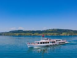 Scenic boat cruising on Lake Morat, surrounded by lush green hills under a clear blue sky near Hotel des Innovations