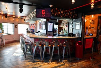 A stylish bar interior featuring a wooden floor, wall decor, stools at the counter, and large windows, at The Gaythering