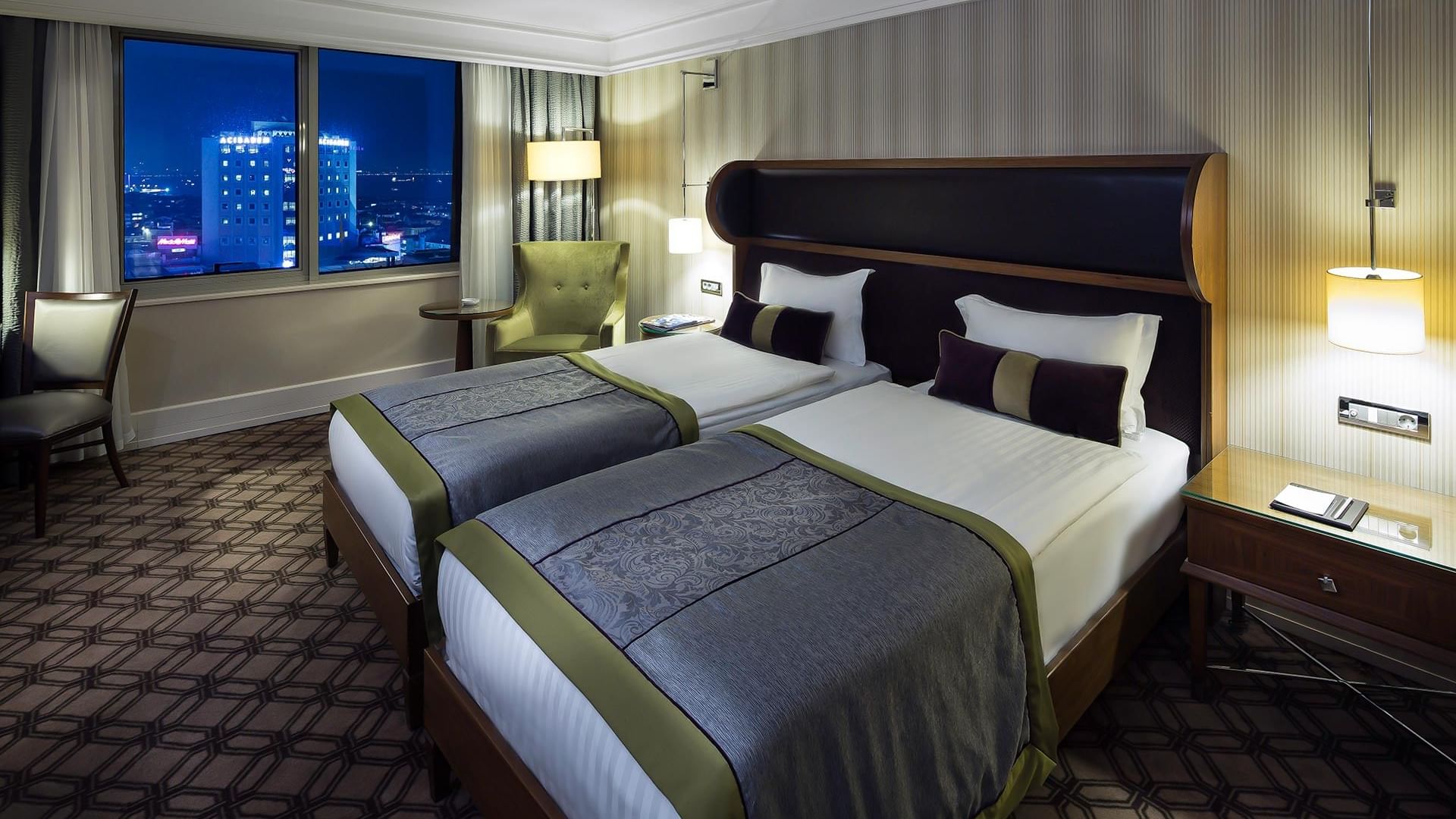 A hotel room with two beds, pillows, nightstands with lamps, a chair, and large windows offering a cityscape view at night in Deluxe Room at Titanic Port Bakırköy