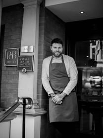 Smiling chef Tom Brown standing in front of restaurant entrance beside wall plaques at Tom Brown at The Capital