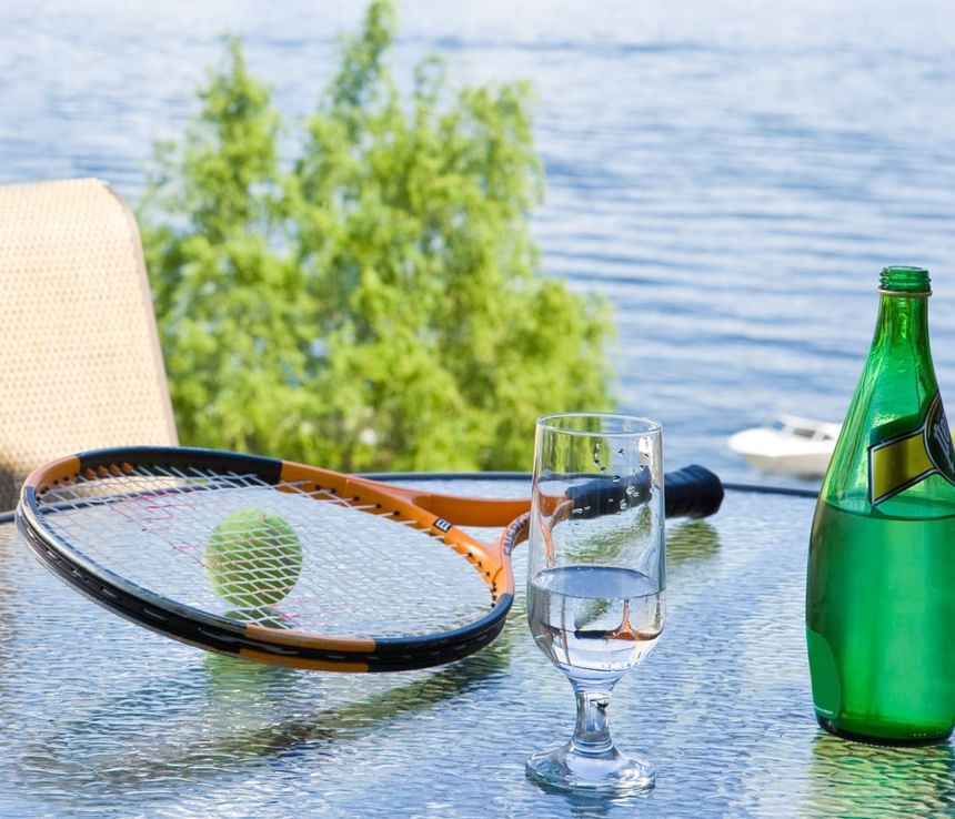 A tennis racket, ball, water glass, and green bottle on a glass table by a lake.