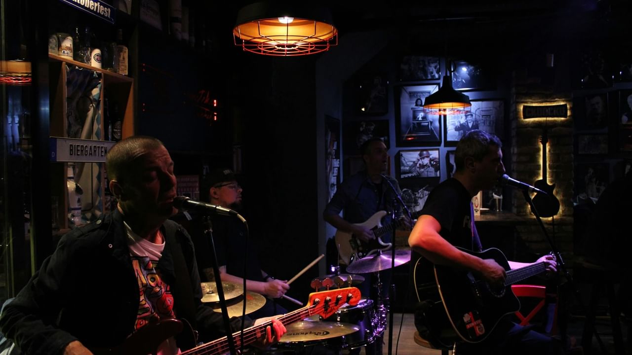 Two men play guitars and sing into microphones while a drummer plays in a dimly lit bar.
