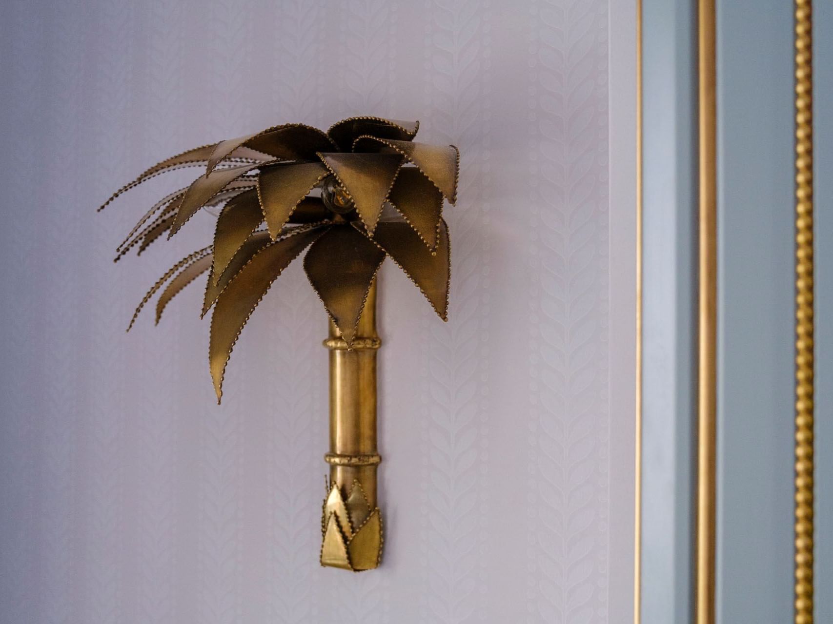 A gold palm tree wall lamp with bamboo stem is mounted on a textured white wall next to a gold-bordered doorway.