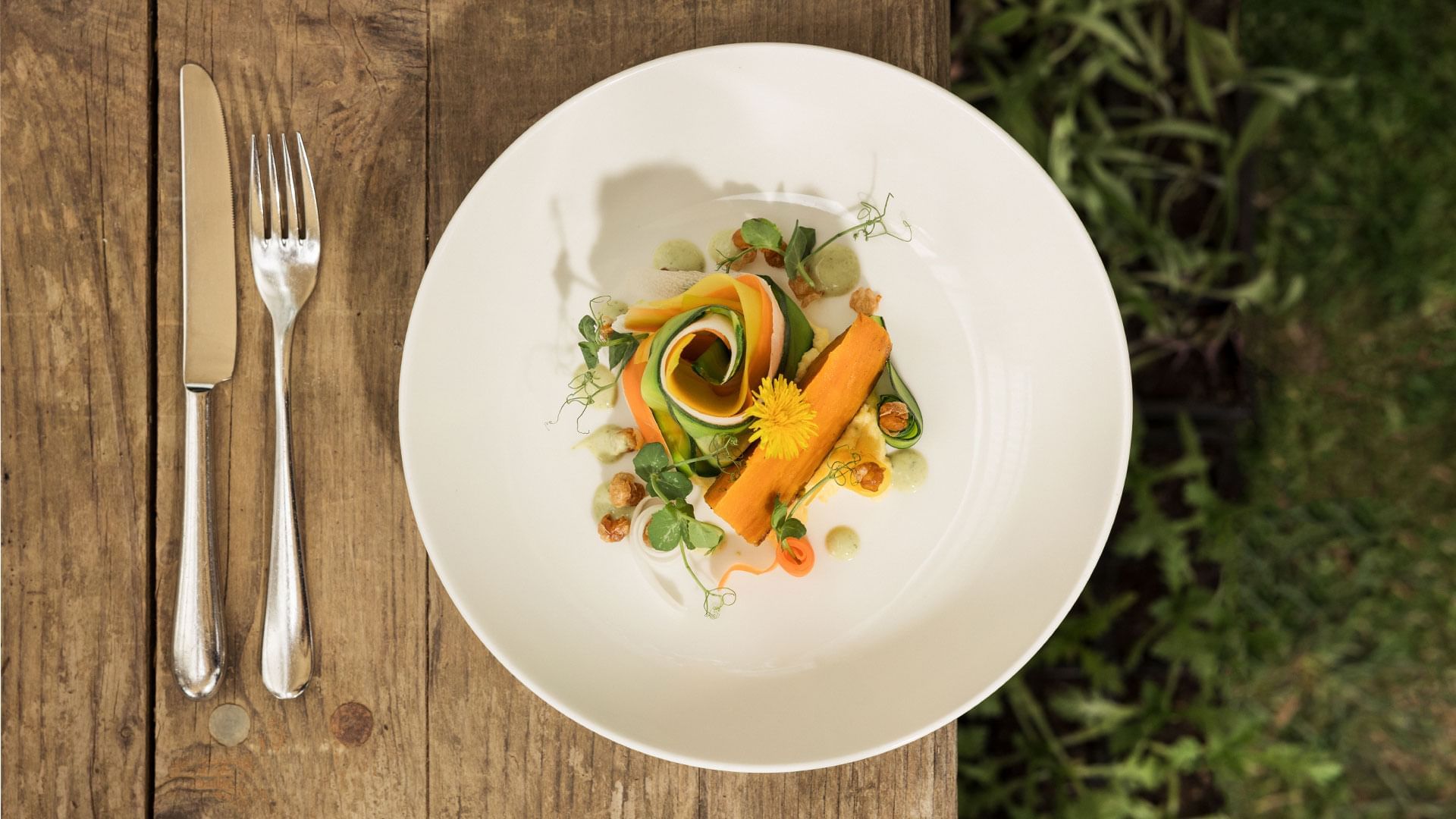 White plate with vegetable salad, knife, and fork on a wooden table with greenery in the background.