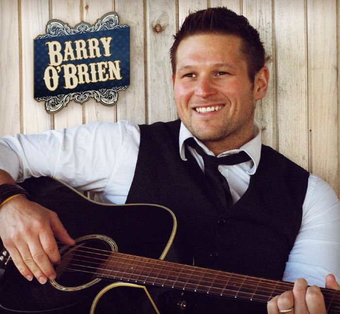Smiling Barry O\'Brien with guitar beside vintage name sign for show at Rodd Brudenell River Resort