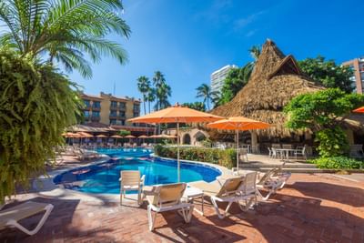 Resort with pool, lounge chairs, umbrellas, thatched-roof hut, and buildings under a clear blue sky.