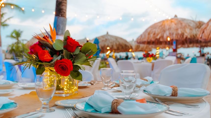 Set dining table with floral centerpiece, blue napkins, and glassware on a beachside venue with string lights and umbrellas.