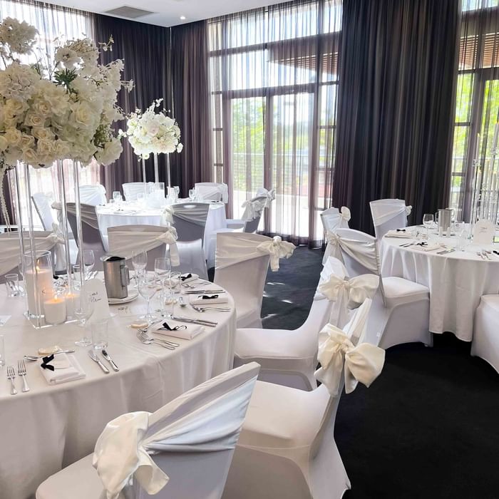 White chairs and tables in a banquet hall with floral centerpieces and curtains.