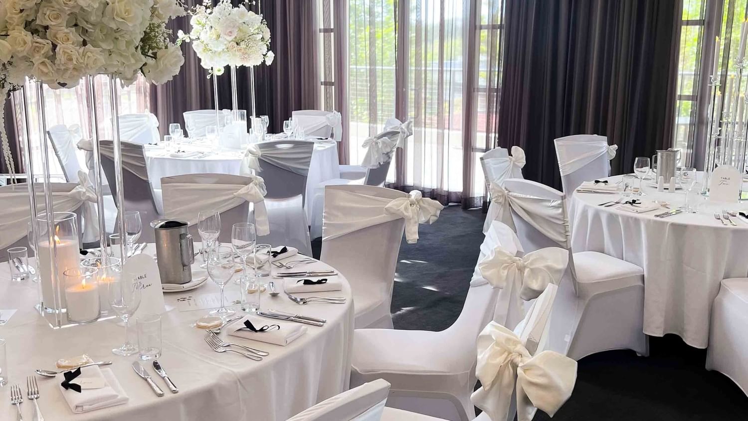 White chairs and tables in a banquet hall with floral centerpieces and curtains.