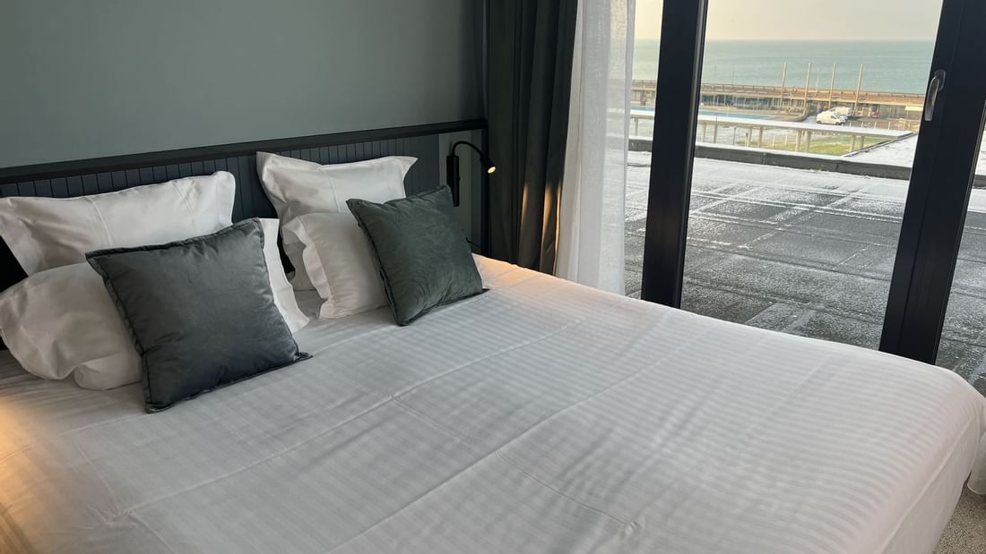 Lit blanc avec oreillers, vue sur la mer par une grande fenêtre.