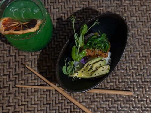 Cocktail and a salmon poke bowl with sliced avocado on a woven placemat in Tortugas restaurant at Hacienda del Mar Los Cabos