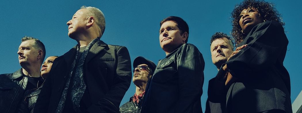 Band members in dark clothing pose against a clear blue sky for Simple Minds – V Tour 2027.