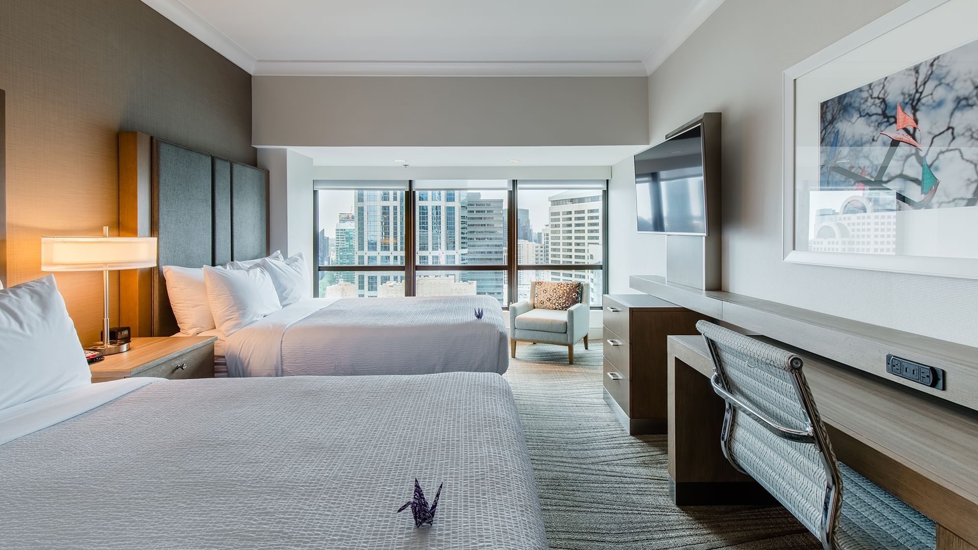 A hotel room with two beds, a desk, chair, and TV, with a city view out the window.