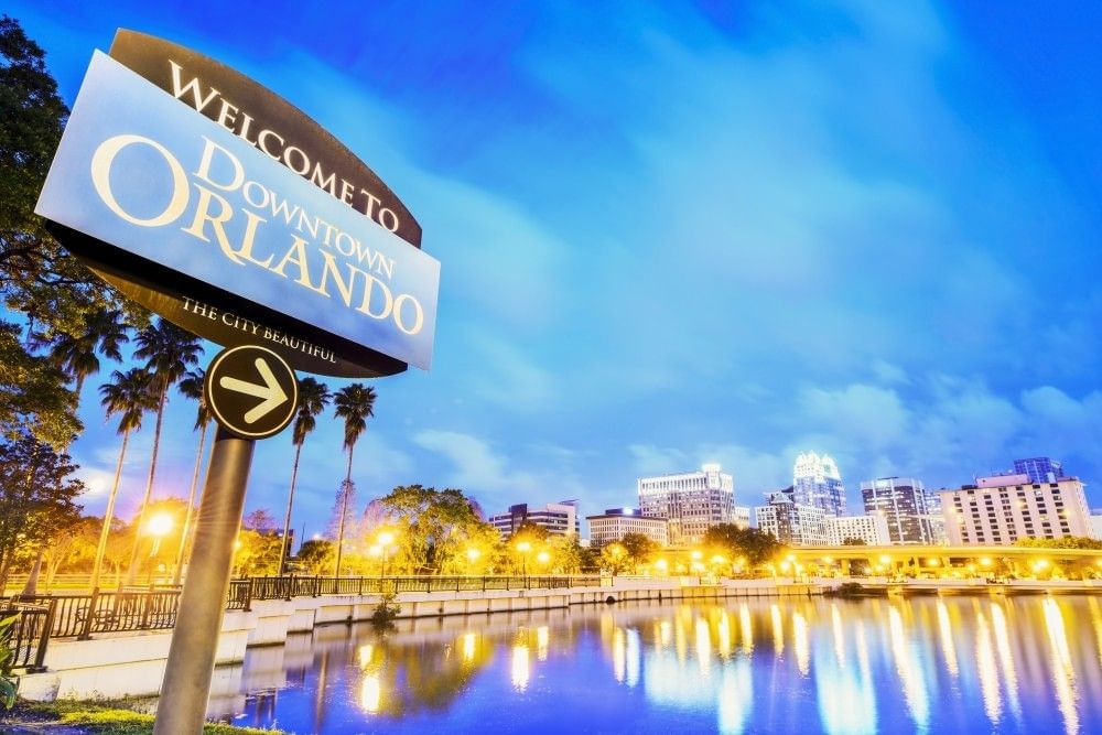 A lake reflecting the building and lights of Downtown Orlando at twilight, and a sign in the foreground that reads Welcome to Downtown Orlando.