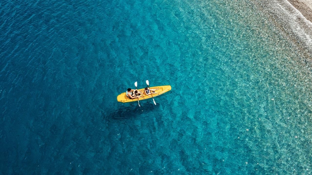 Two people in a yellow kayak with paddles on a blue ocean near a beach.