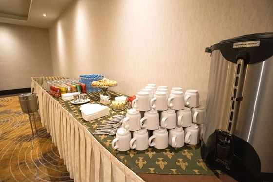 Estación de café con tazas apiladas, urna plateada, bebidas enlatadas y pasteles sobre una mesa cubierta en Araiza Calafia