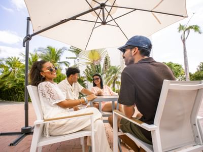 Four guests relaxing on white lounge chairs under a beige umbrella surrounded by tropical palms at Playa Blanca Beach Resort
