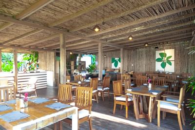 Gigi Beach dining with wooden tables and sunlight through a thatched roof at Le Cameleon Boutique Hotel