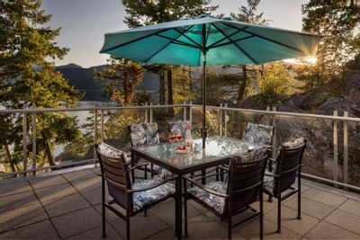Outdoor patio with dining table and chairs under a teal umbrella overlooking a lake and forest.